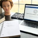 A photo of a desk with a laptop open to a state board of cosmetology license application portal, a "Standard Cosmetology" textbook, a notebook with study notes, and a mannequin head.