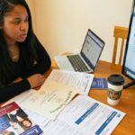 A young woman sits at a table with a High School Equivalency diploma and other documents, looking concerned while navigating an online school application portal