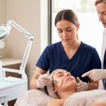 A mentor supervises an esthetician student applying a facial treatment with a brush to a client in a professional medical spa environment.