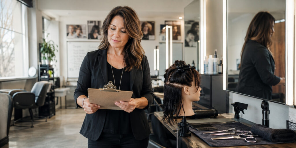 Experienced beauty instructor reviewing a clipboard beside a mannequin head and salon tools in a cosmetology classroom.
