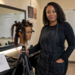 Experienced beauty instructor standing in a cosmetology classroom beside a sectioned mannequin head, open lesson plan, styling tools, and instructor training notes.