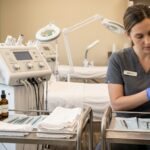 A professional skincare practitioner in gray scrubs reviews a skin analysis chart next to advanced facial machines and a treatment tray with tools.