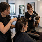 Cosmetology student carefully sectioning a client’s hair during hands-on training while other students practice hair coloring and nail services in the background.