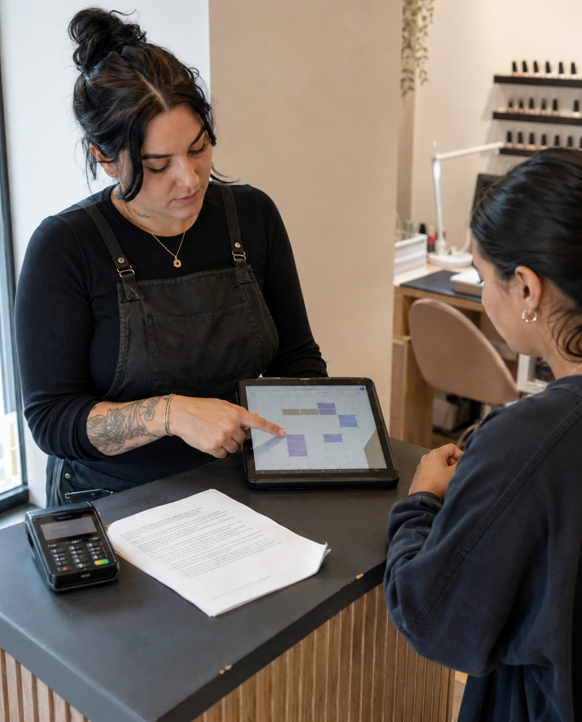 Nail technician explaining appointment rules to a client using a tablet calendar beside a policy sheet and payment terminal at a salon reception counter.