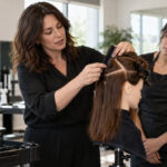 Cosmetology educator demonstrates comb placement and precise sectioning on a brown-haired mannequin while three adult students observe around a classroom workstation.