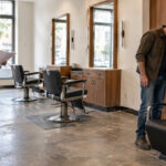 Prospective barbershop owner holds a floor plan while a contractor examines a shampoo basin beside empty barber chairs and existing workstations.